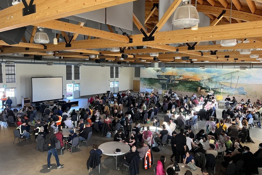 The staff of Fort Edmonton Park is sitting at round tables at the Blatchford Hangar on 1920s Street. They are looking at a stage and screen as part of the annual Safety Day.