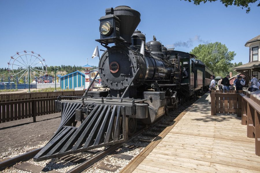 The 1919 Baldwin Locomotive 107 sits at the Fort Edmonton Park front-entry train station. Guests are observing the black and green train. In the background, you can see the 1920s Midway with a Ferris wheel.