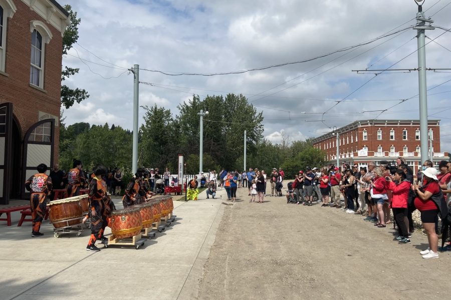 Seven drummers play on 1905 Street at Fort Edmonton Park in front of the fire hall. A crowd watches them from the road.