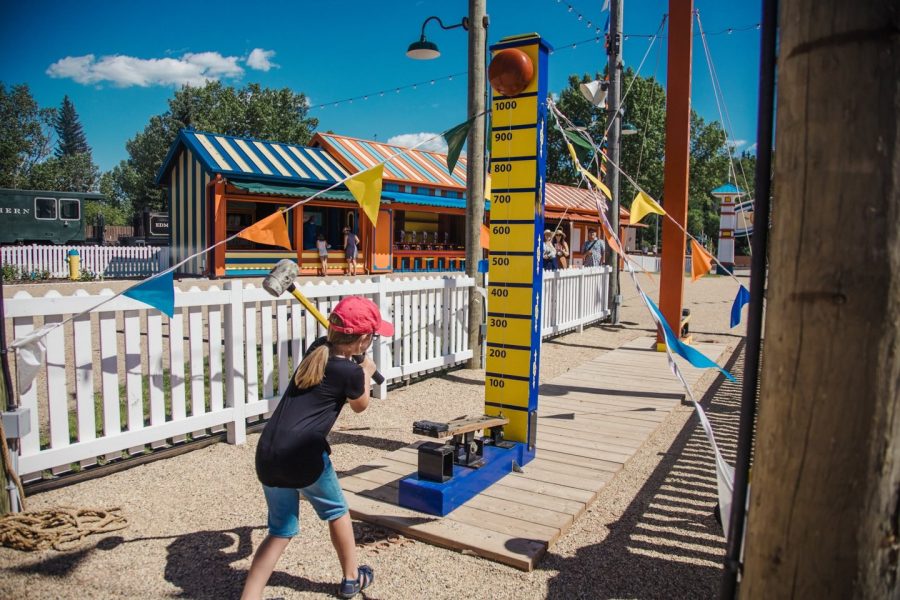 Midway-Bell-Game A child tries their hand at the sledgehammer and bell game. In the background, there are more games with other guests planning them.