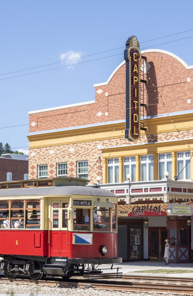 A red streetcar passes in front of the Capitol Theatre at Fort Edmonton Park. There is a 1920s interpreter in front of the theatre.