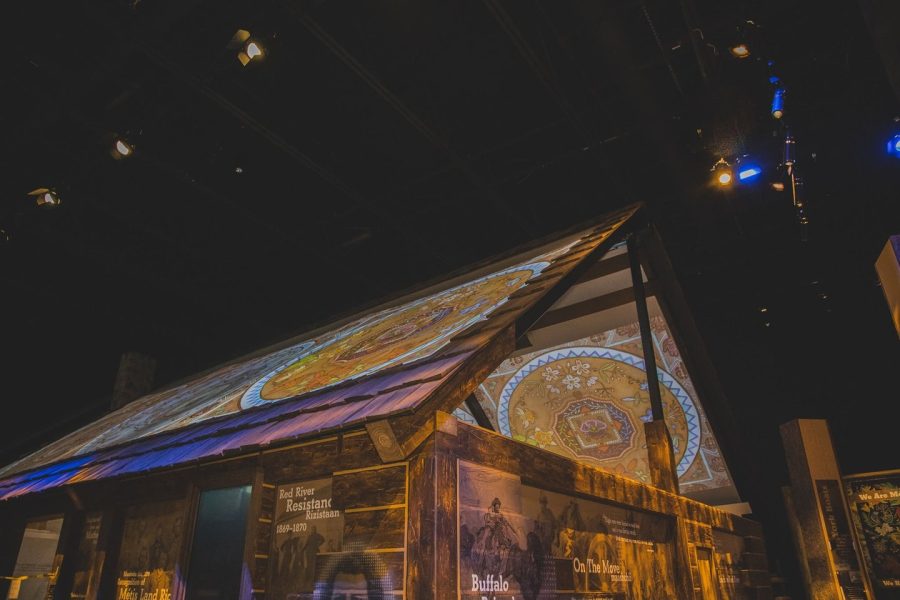 A photograph of the Métis Cabin inside the Indigenous Peoples Experience at Fort Edmonton Park. The roof has projection screens with beaded work, and the outside walls have images that tell Métis stories.